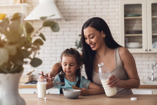 Photo a young happy mother prepares breakfast for her little daughter by adding milk and muesli to a plate while sitting at a table in the kitchen at home