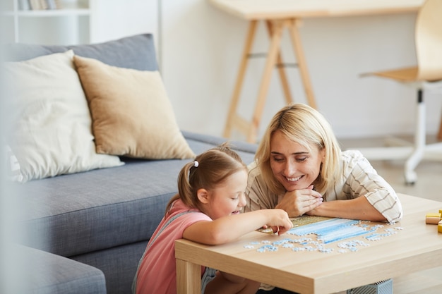 Young happy mother teaching her daughter with down syndrome collecting puzzles at the table in the living room