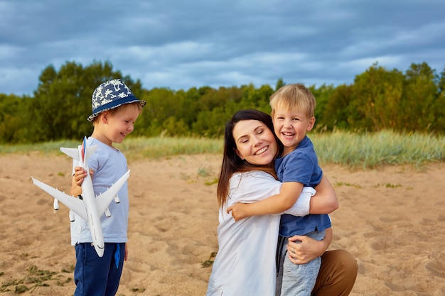 Молодая счастливая мама с двумя мальчиками играет на песке возле леса, в руках у одного из них модель гражданского самолета и шляпа на голове