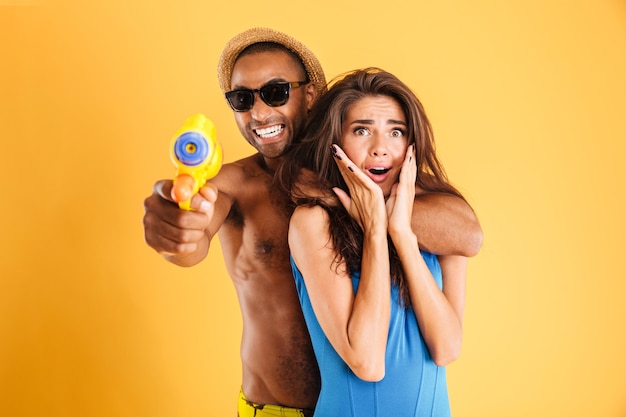 Young happy multiracial couple having fun playing with water guns isolated on the orange wall