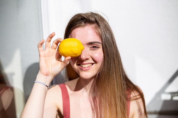 Young happy pretty female in casual summer dress sitting on terrace and covering eye with ripe lemon