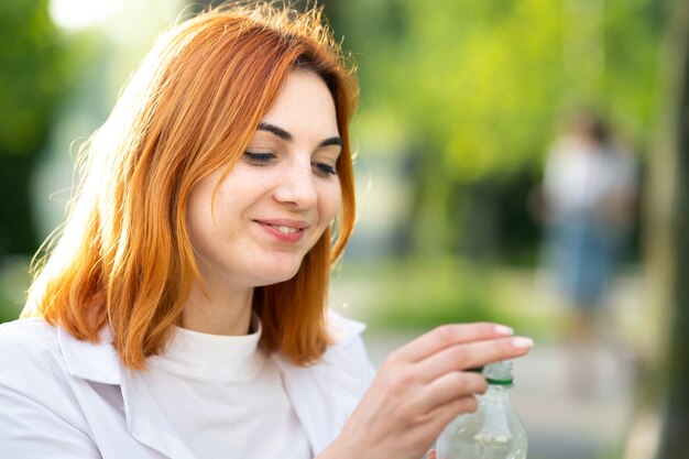 夏の公園で彼女の手で水のボトルを保持している若い幸せな赤毛の女の子。