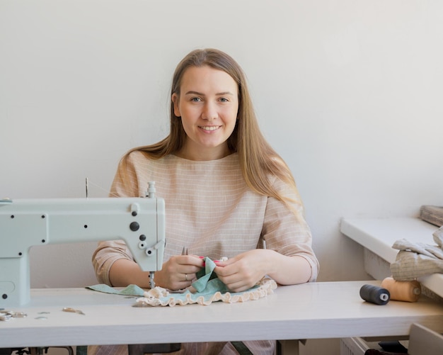Фото Молодая счастливая швея шит одежду в собственной мастерской улыбается в камеру женщина-владелец мастерской