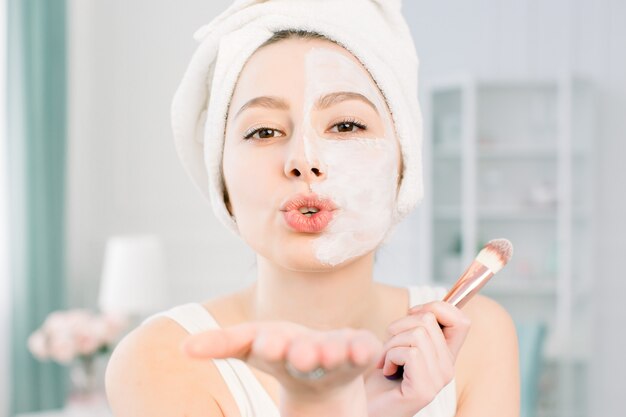 Young happy and smiling beautiful woman applying clay facial mask to half the face and sending an air kiss