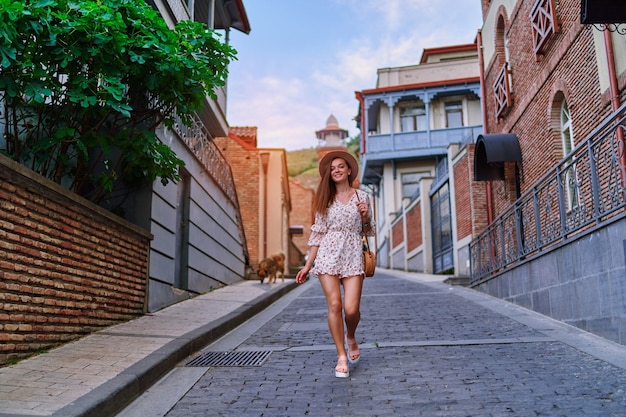 Young happy smiling cute beautiful brunette traveler girl with long legs wearing hat, round straw bag and short jumpsuit walking alone around the city at sunny summer day