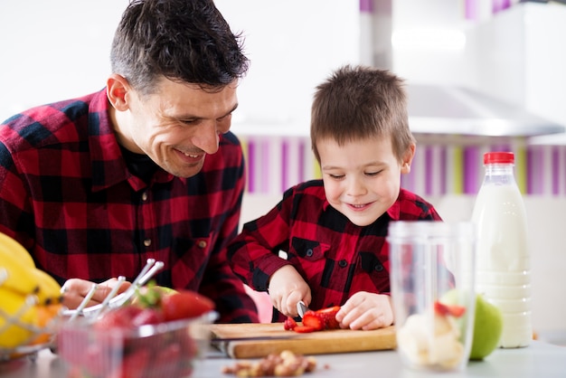 若い幸せな笑顔の男性の子供は、父親と一緒にキッチンカウンターでイチゴを切るときにナイフを使用する方法を考えています。