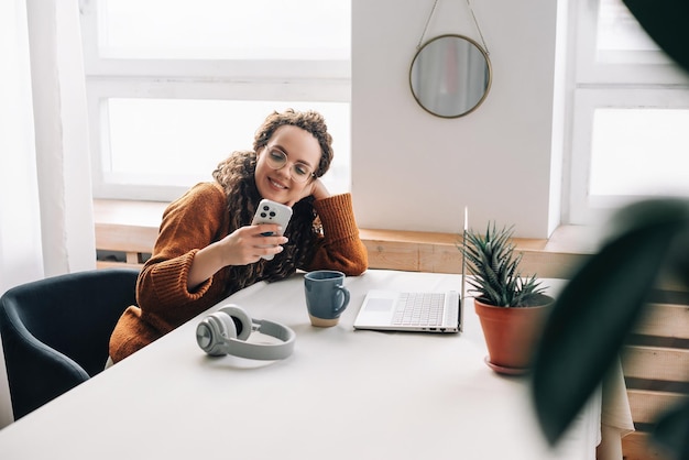 Young happy smiling pretty woman sitting on a chair holding smartphone using cell phone mobile