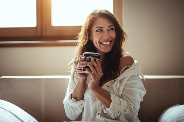 웃고 있는 젊은 여성이 잠에서 깨어난 후 방에 앉아 모닝 커피를 마시고 있습니다.