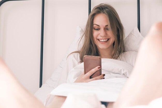 Giovane donna sorridente felice che usa il suo telefono mentre si trova a letto la ragazza del risveglio mattutino legge la sostanza appiccicosa