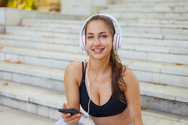 Young happy sport woman in sportswear use smartphone and listens to music in headphones while sitting on stairs on bright sunny day