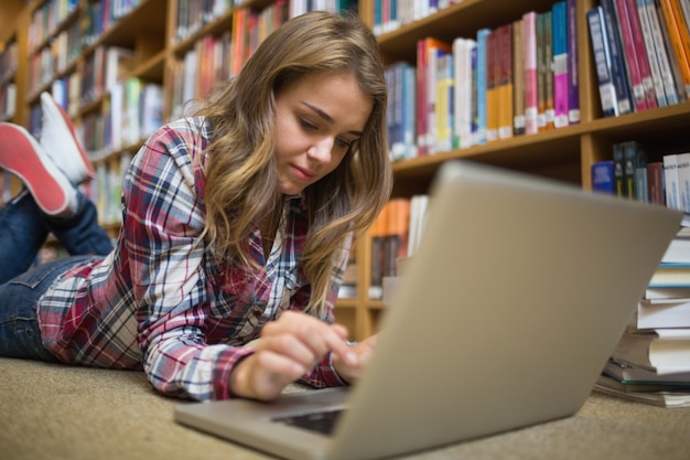 Молодой счастливый студент, лежа на полу библиотеки, используя ноутбук