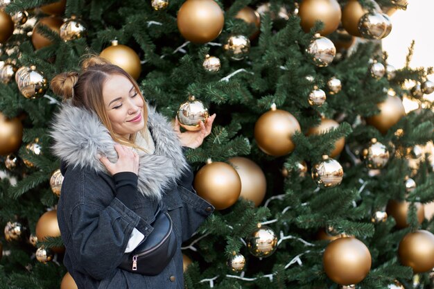 Young happy stylish woman with closed eyes posing on the background of the christmas tree with golden balls.