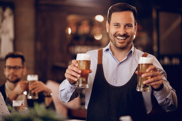 Young happy waiter holding two glasses of beer and looking at camera while working in a bar