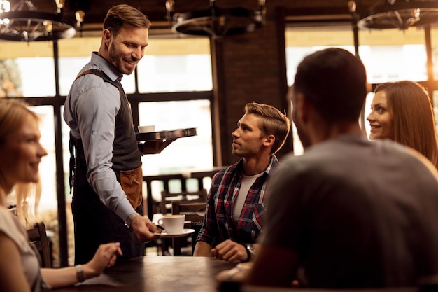Young happy waiter serving coffee to group of friends in coffee shop