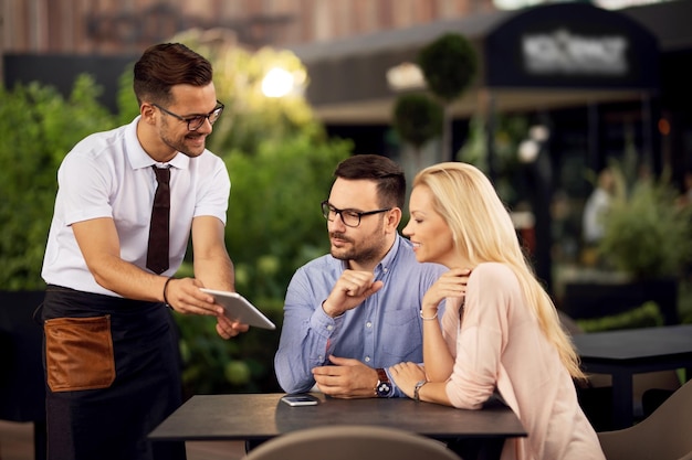 Young happy waiter serving a couple and showing them menu on digital tablet in a restaurant