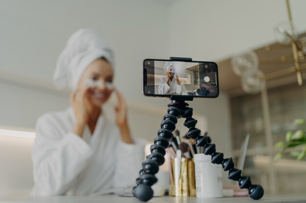 Young happy woman beauty blogger in bathrobe recording video tutorial for skin care blog about using cosmetic under eye patches pads to deal with wrinkles and dark circles, focus on smartphone screen