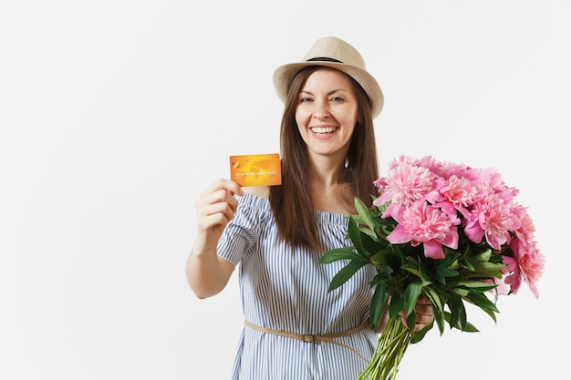 Young happy woman in blue dress, hat holding credit bank card, money, bouquet of beautiful pink peonies flowers isolated on white background. Business, delivery, online shopping concept. Copy space.