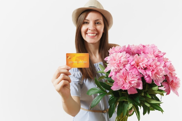 Young happy woman in blue dress, hat holding credit bank card, money, bouquet of beautiful pink peonies flowers isolated on white background. Business, delivery, online shopping concept. Copy space.