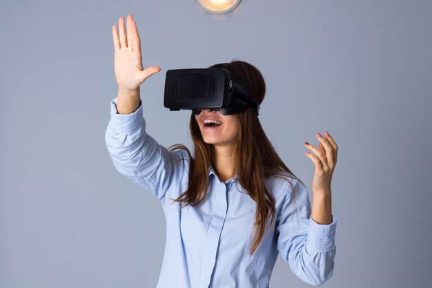 Young happy woman in blue shirt using VR glasses on grey background in studio