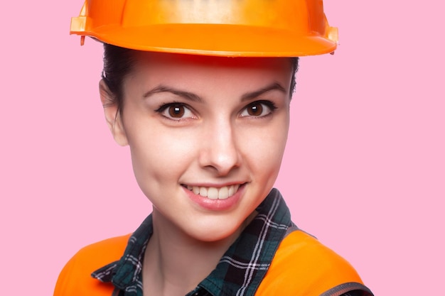 young happy woman builder in hardhat and orange vest pink background