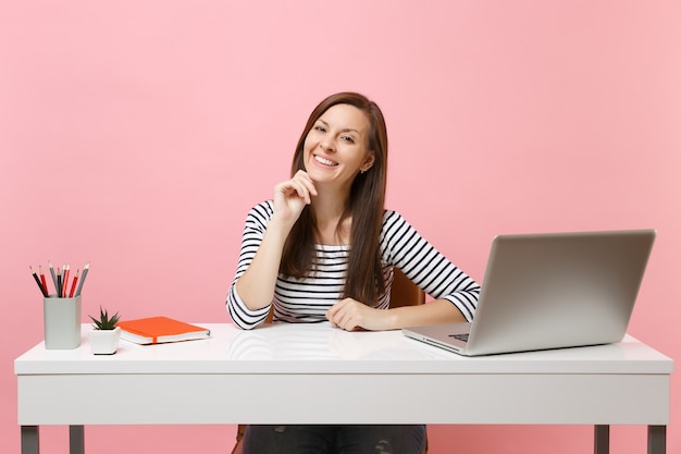 Giovane donna felice in abiti casual con il mento appoggiato sulla mano si siede e lavora alla scrivania bianca con un computer portatile contemporaneo