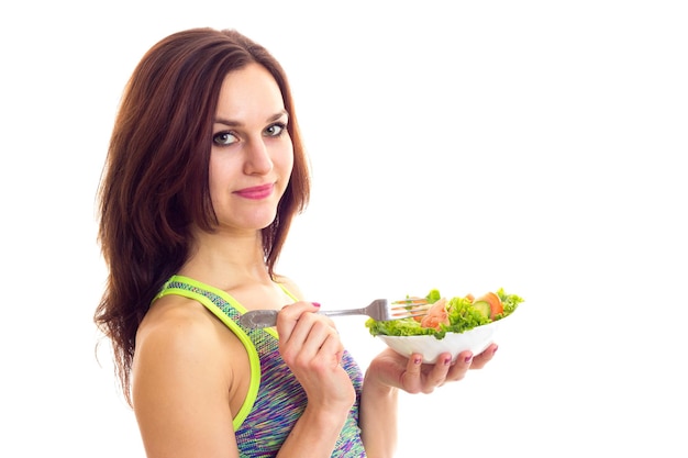 Молодая счастливая женщина в цветном спортивном топе держит вилку и белую тарелку с зеленым салатом и помидорами