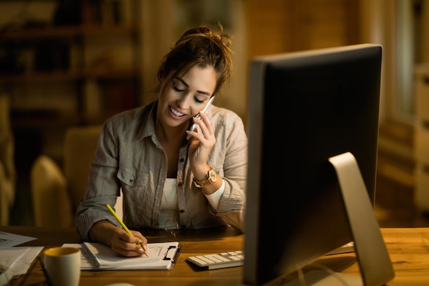 Young happy woman communicating on mobile phone and taking notes while working late at home