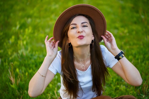 모자를 쓴 젊은 행복한 여성은 공원의 푸른 잔디밭에 앉아 있습니다. 밝고 화창한 여름날 얼굴에 미소를 머금은 유럽풍 소녀
