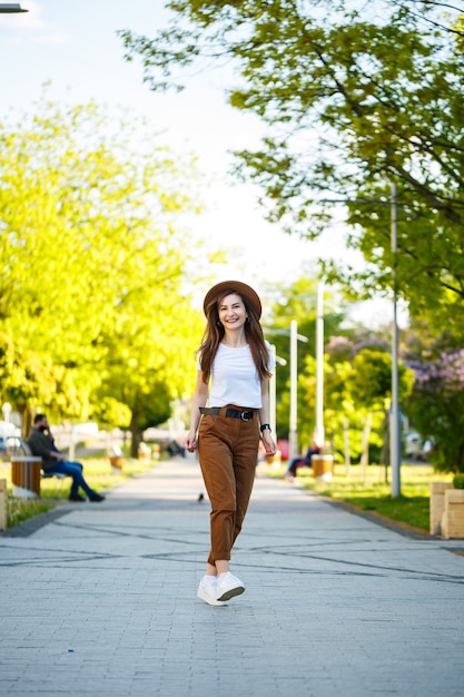 帽子をかぶった若い幸せな女性が公園の路地を歩いています。明るい晴れた夏の日に彼女の顔に笑顔でヨーロッパの外観の女の子