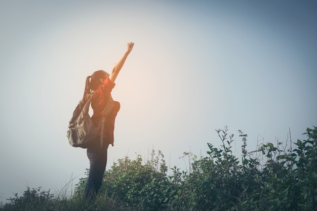 Young happy woman hiker with backpack standing on mountain peak with open arms