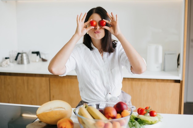 young happy woman holding cherry tomato at eyes and showing kiss on background of modern white kitchen healthy food concept home cooking salad ingredients