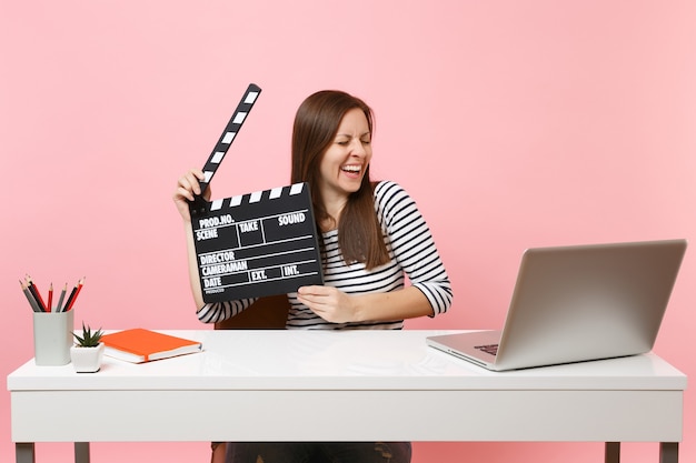 Young happy woman holding classic black film making clapperboard working on project while sit at office with laptop isolated on pastel pink background. Achievement business career concept. Copy space.