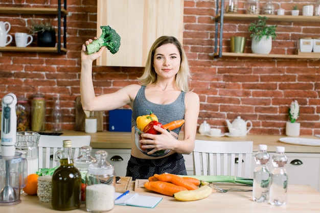 Young and happy woman holding glass bowl with fresh vegetables in left hand and broccoli in right hand while standing on the table with healthy food in modern kitchen. Healthy food concept