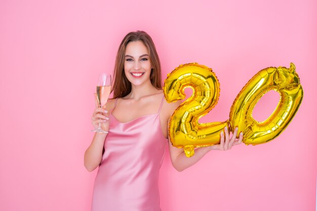 Young happy woman holds gold foil balloon and glass of champagne on pink background birthday party