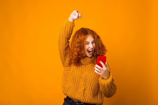 Young happy woman is celebrating, looking at the phone with an arm up on orange wall.