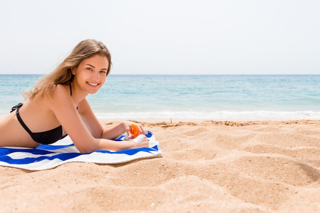 Young happy woman is resting on the towel near the sea and protects her skin on the hand with sunscreen from the spray.