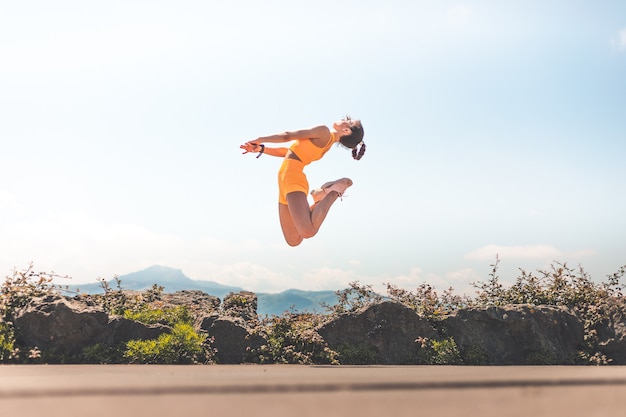 青い空を背景に設定されたオレンジ色のスポーツでジャンプする若い幸せな女性