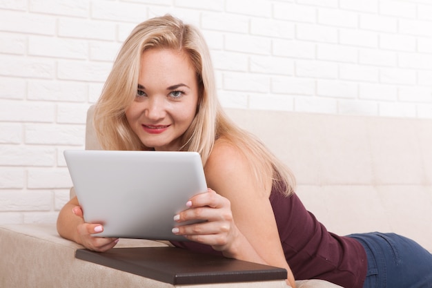 Young happy woman lying on the sofa with pillows and using tablet computer at home