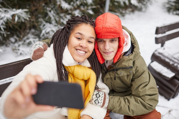 Giovane donna felice che propone alla macchina fotografica del suo telefono cellulare insieme all'uomo mentre sono seduti su una panchina in inverno