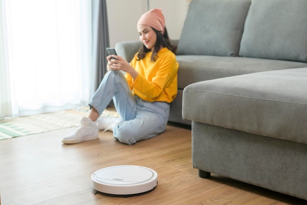 Молодая счастливая женщина расслабляется и пользуется смартфоном в гостиной, пока работает робот-пылесос