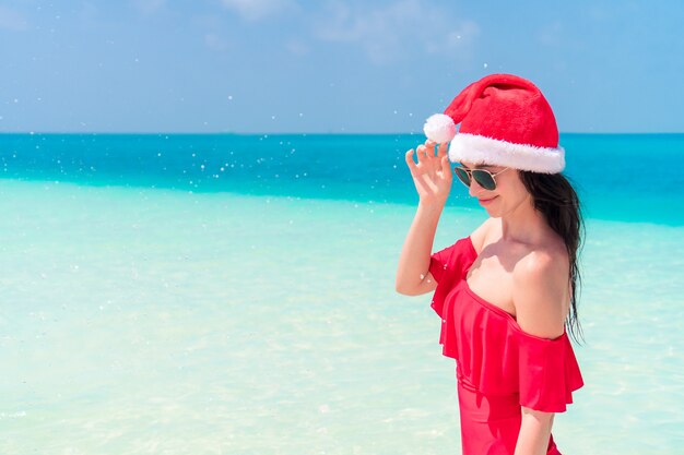 Young happy woman in Santa hat in swimsuit on white beach on Xmas holidays