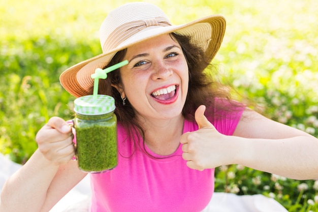 ピクニックで緑のスムージーと親指を見せて若い幸せな女性。健康食品、デトックス、ダイエットのコンセプト