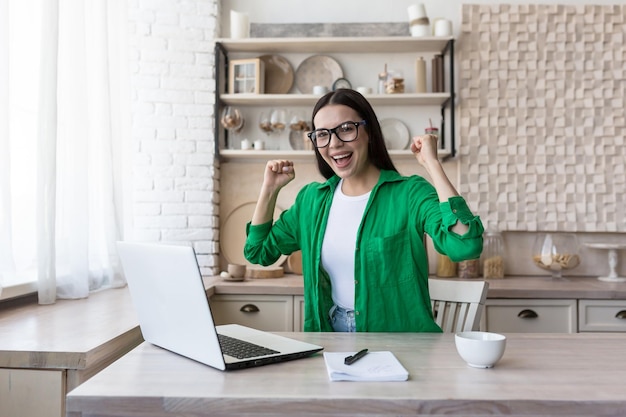 A young happy woman sits at home in front of a laptop and rejoices raising her hands up celebrating