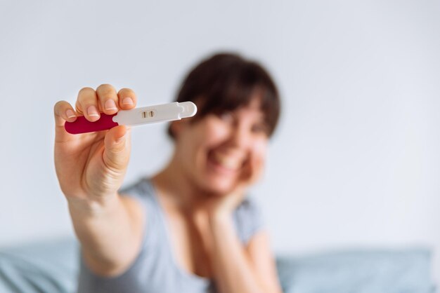 Young happy woman sitting on bed looking on positive pregnancy test blurred background