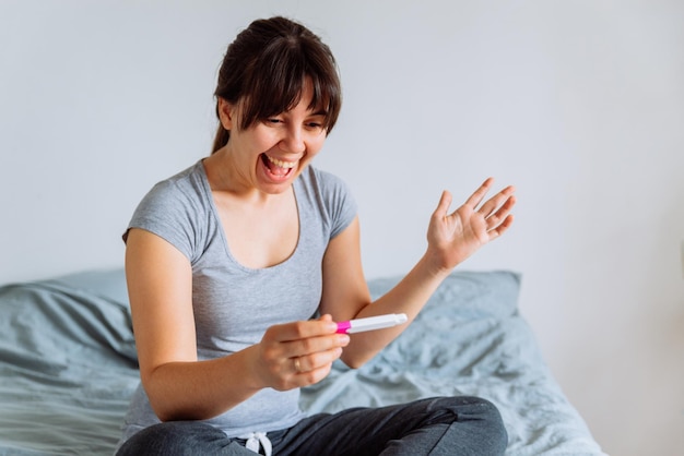 Young happy woman sitting on bed looking on positive pregnancy test