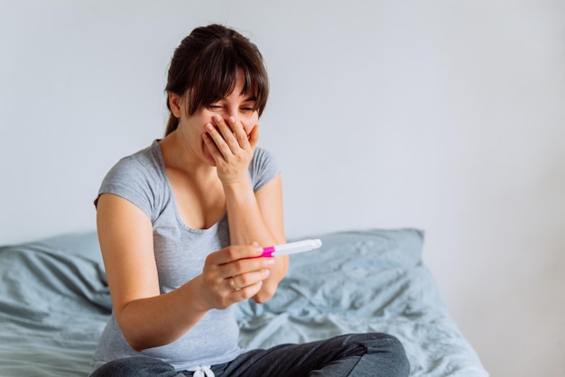 Young happy woman sitting on bed looking on positive pregnancy test