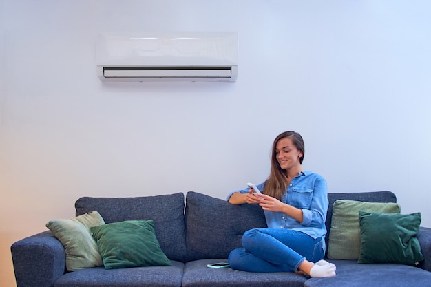 Young happy woman sitting on couch under air conditioner and adjusting comfort temperature with remote control at modern home