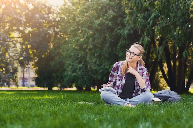 Young happy woman sitting with notebook on grass. Woman studying outdoors with copy-book. Education, inspiration and remote working concept.