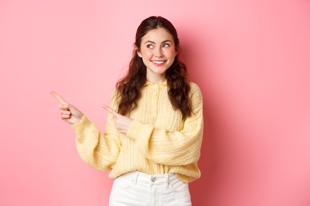 Young happy woman smiling, pointing and looking aside at left copyspace, showing advertisement, standing against pink spring wall.