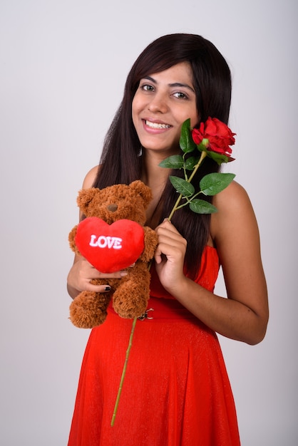 Young happy woman smiling while holding red rose and teddy bear with heart and love sign ready for Valentine's day   .